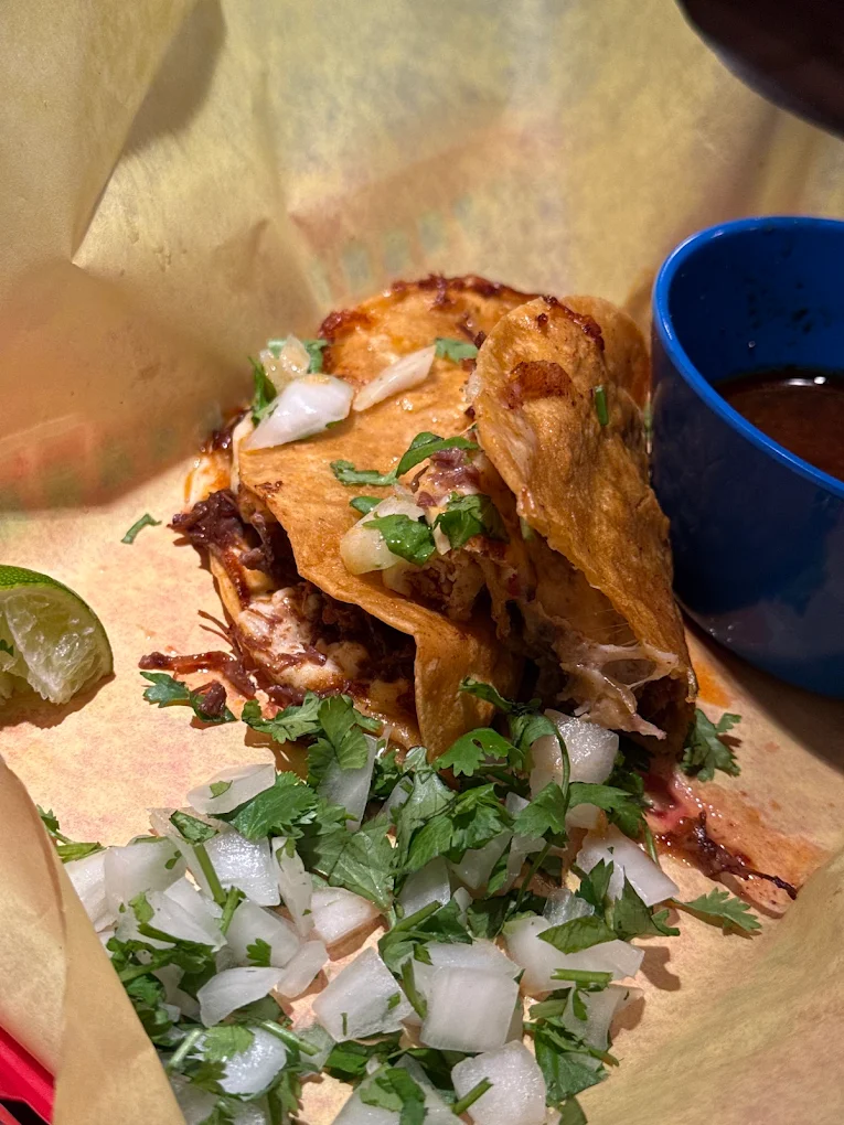 Griddled quesabirria tacos with melted cheese pulling apart, topped with onion and cilantro, served with a blue cup of consommé and a lime wedge.