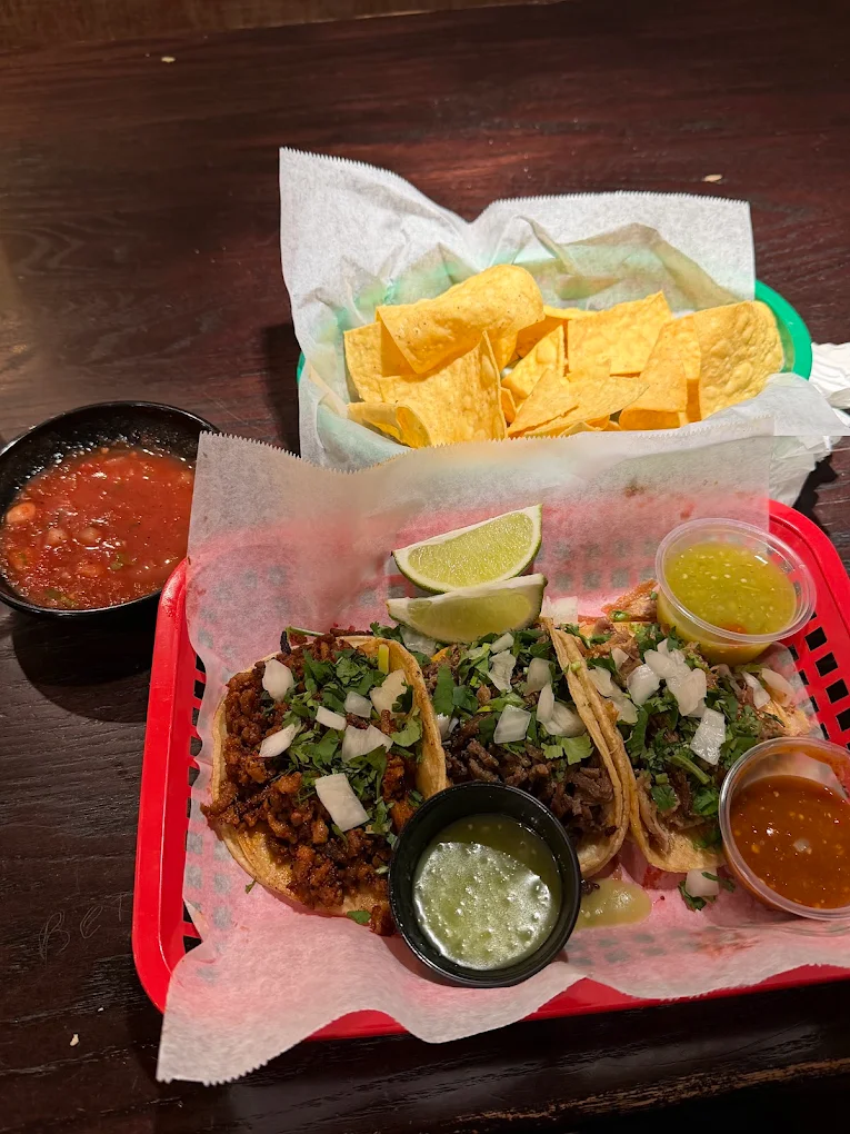 Three corn tortilla tacos in a red basket with onion and cilantro, lime wedges, a basket of tortilla chips, and cups of red and green salsa.