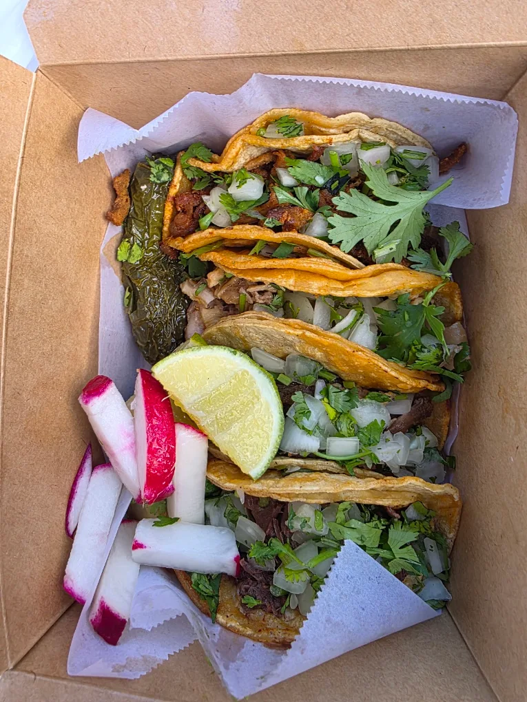 A takeout box of tacos topped with grilled nopales and onion, with sliced radish and a lime wedge alongside.
