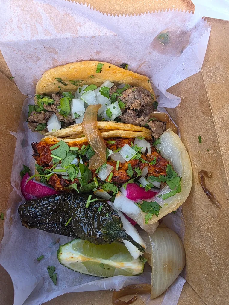 A takeout tray of tacos with cilantro, onion and pickled radish, with grilled onion, a roasted pepper and a lime wedge on the side.
